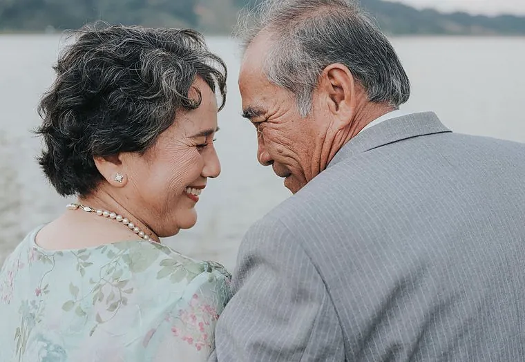An elderly couple sharing a quiet moment by the water, smiling at each other
